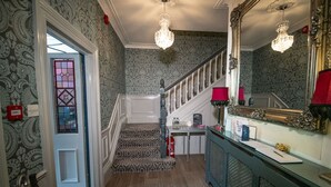 A hallway with patterned wallpaper, a staircase, a desk with a lamp, and a mirror.