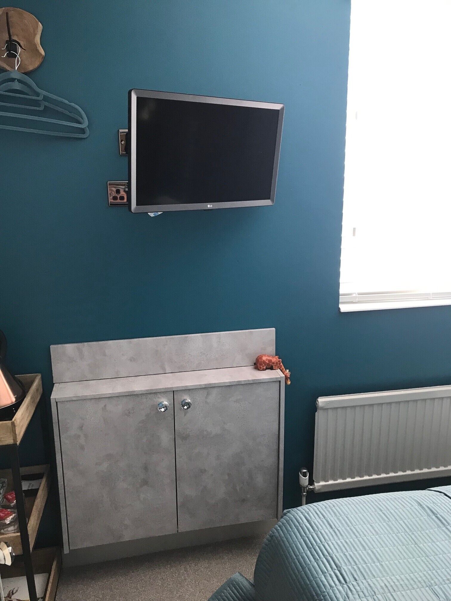 Ein Schlafzimmer mit an der Wand montiertem Fernseher, einem grauen Kleiderschrank, einem Heizkörper und einem Fenster.