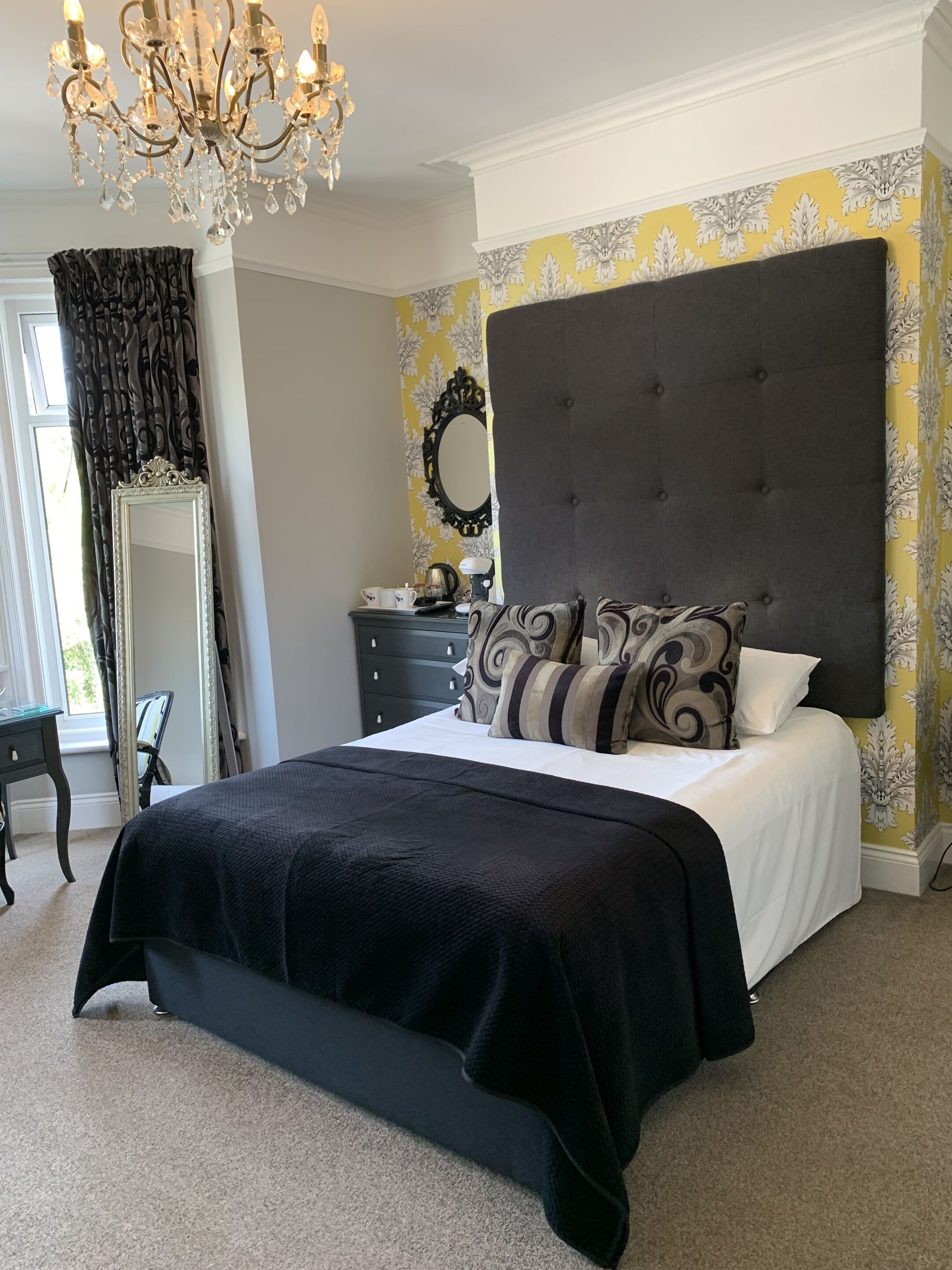 A bedroom with a large bed, a dark headboard, a chandelier, a dresser, and a mirror.