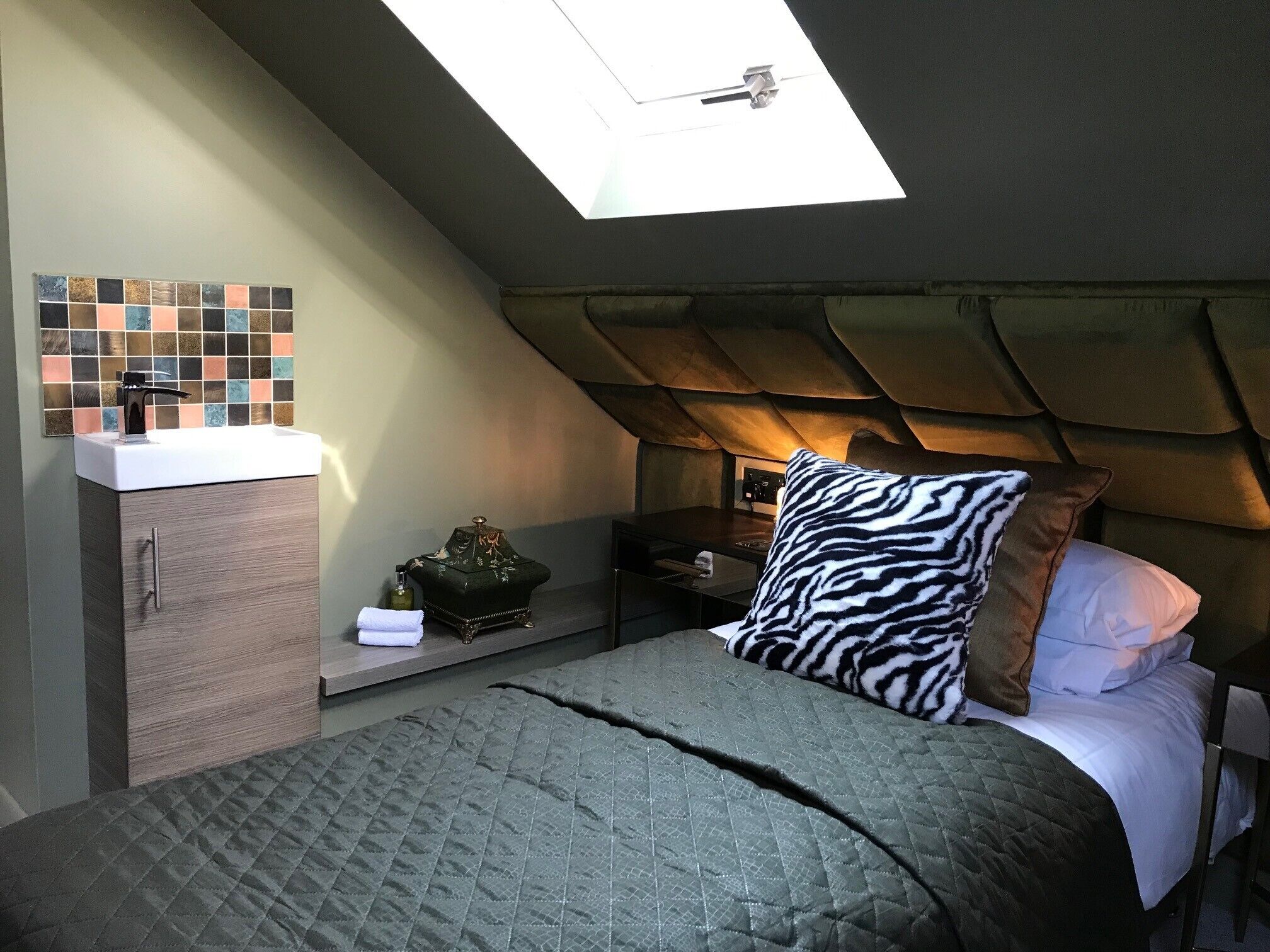 Quarto com claraboia, cama com travesseiro estampado com padrão de zebra, mesa de cabeceira, armário e pia fixa na parede.