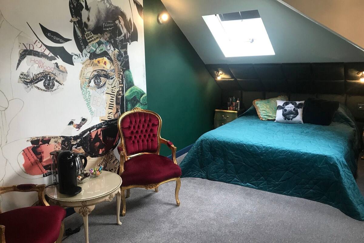 A bedroom with a sloped ceiling, a large mural on the wall, a bed with teal bedding, a red upholstered chair, and a small table with a coffee pot.