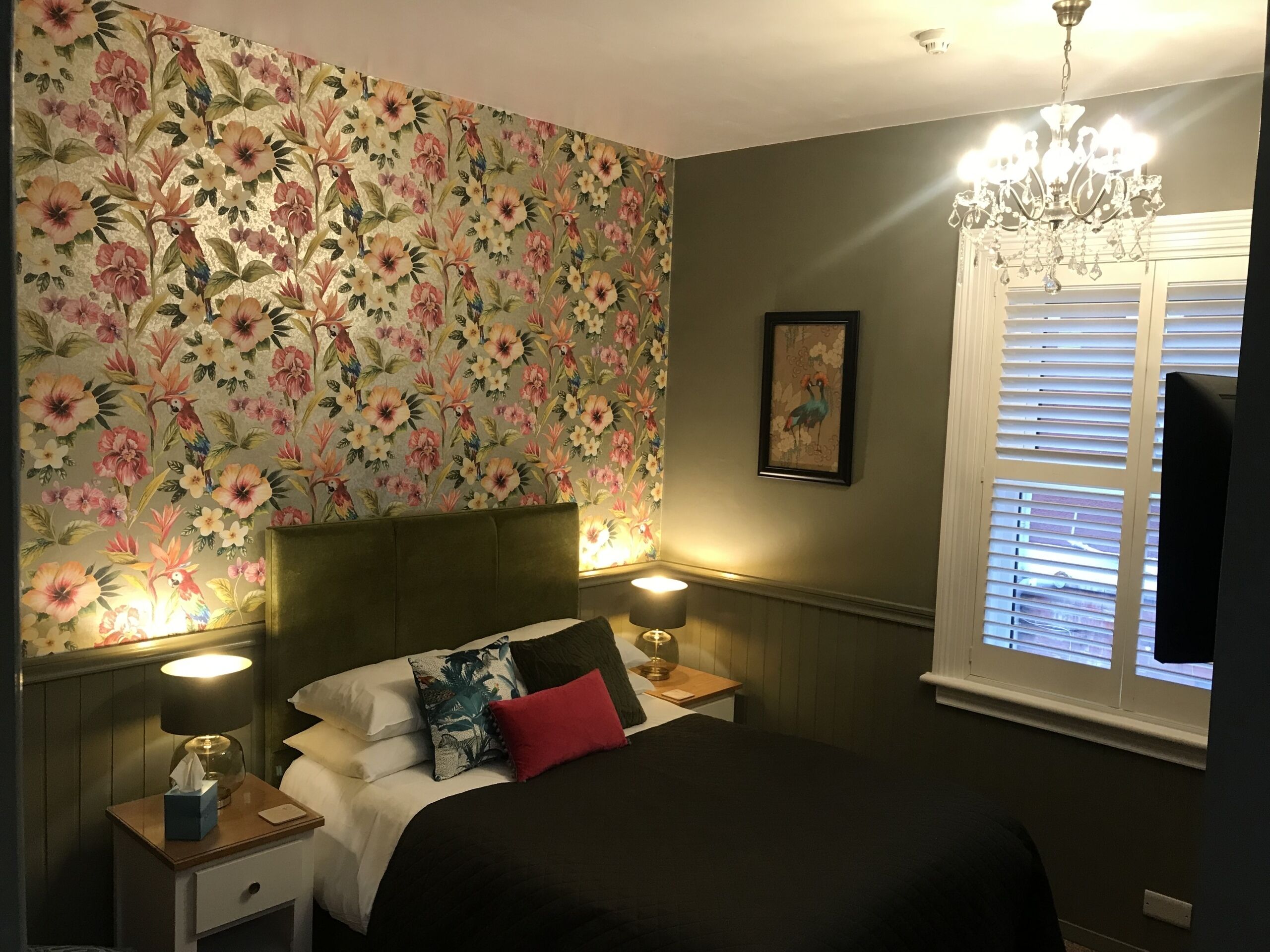 Ein Schlafzimmer mit blumigem Wanddesign, einem Bett mit vielen Kissen, Nachttischlampen, einer Kronleuchter und einem Fenster mit Jalousien.