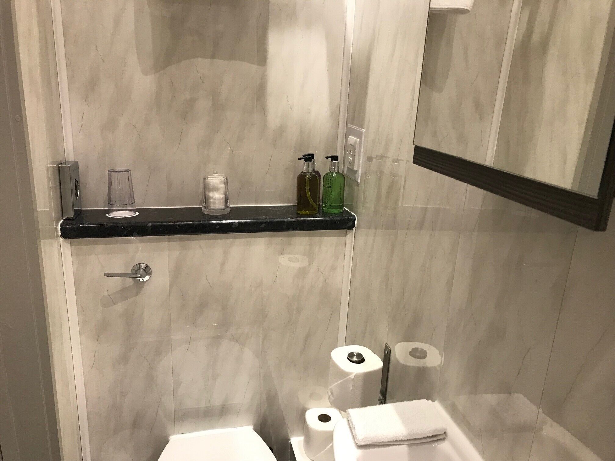 A bathroom with a marble wall, a black countertop, a toilet, a sink, a mirror, and a shelf with toiletries.