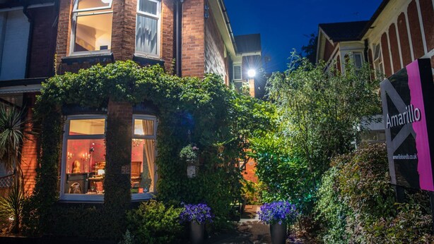 A two-story house with a brick facade, a large window, and a garden with potted plants and flowers.