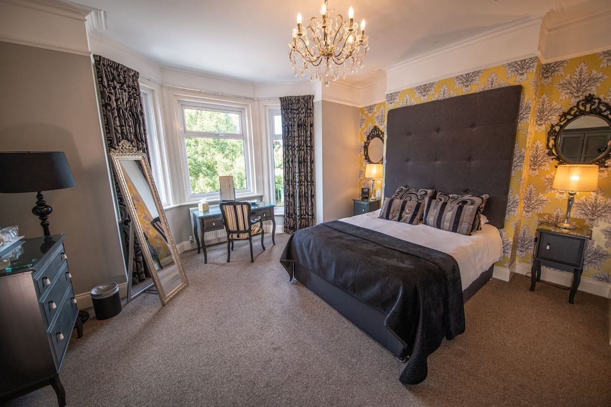 A bedroom with a large bed, a desk, and a chair. There is a chandelier above the bed and a mirror on the wall.