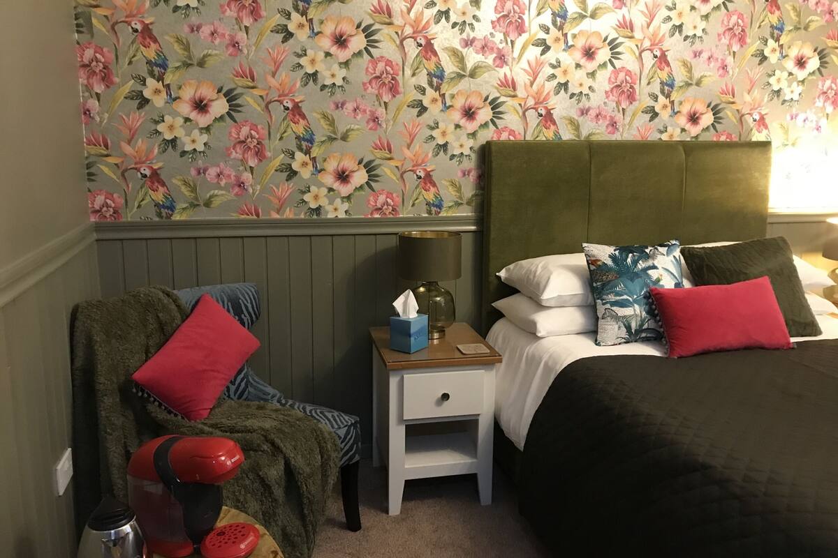 A hotel room with a floral wallpaper, a green upholstered headboard, a bed with multiple pillows, a nightstand with a lamp and a tissue box, and a chair with a red cushion.