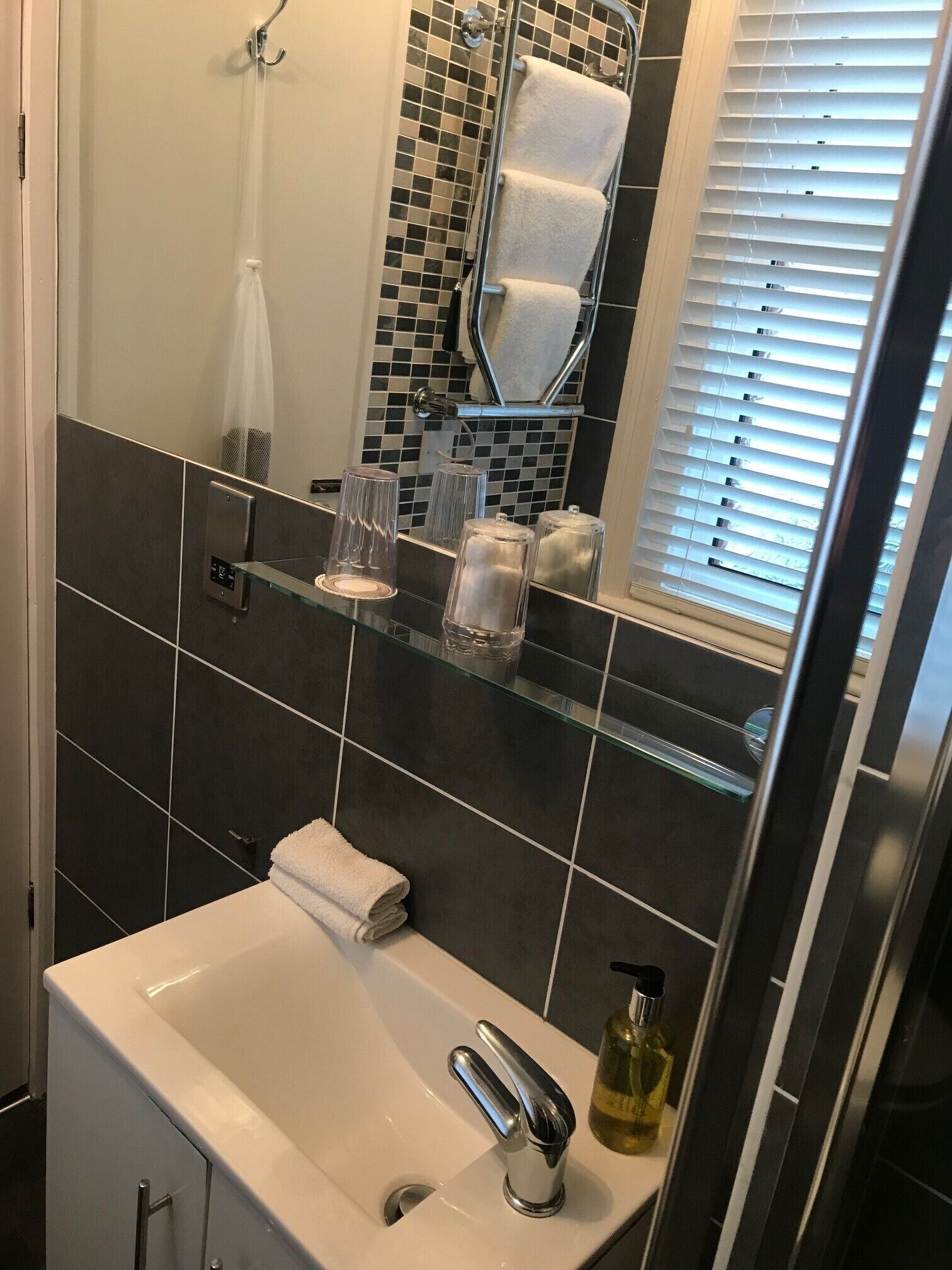 Un baño con lavamanos blanco, repisa de vidrio con artículos de aseo, toallero y zona de ducha con pared azulejada.