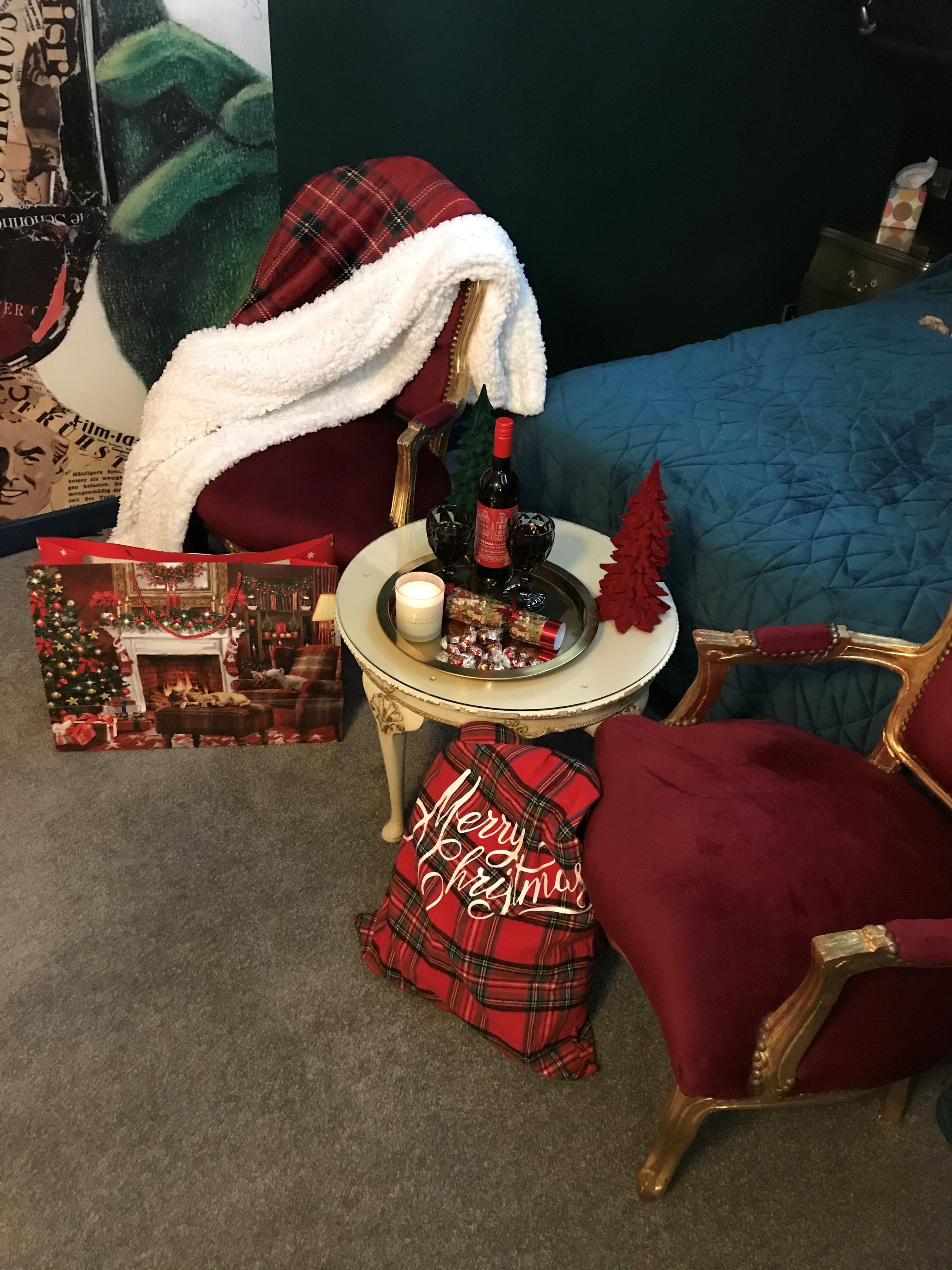 A cozy room with a red plaid chair, a small round table with a candle, a bottle of wine, and a Christmas tree decoration.