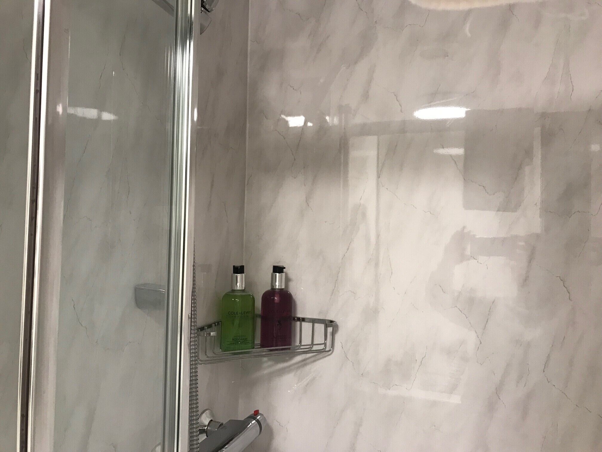 A bathroom with a glass shower enclosure, marble walls, and two shelves holding toiletries.