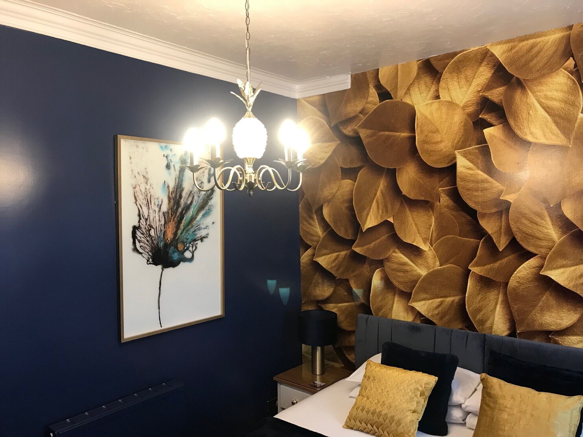 Quarto com parede azul escura, cama com cabeceira estampada em folhas douradas, lustre e uma obra de arte emoldurada.