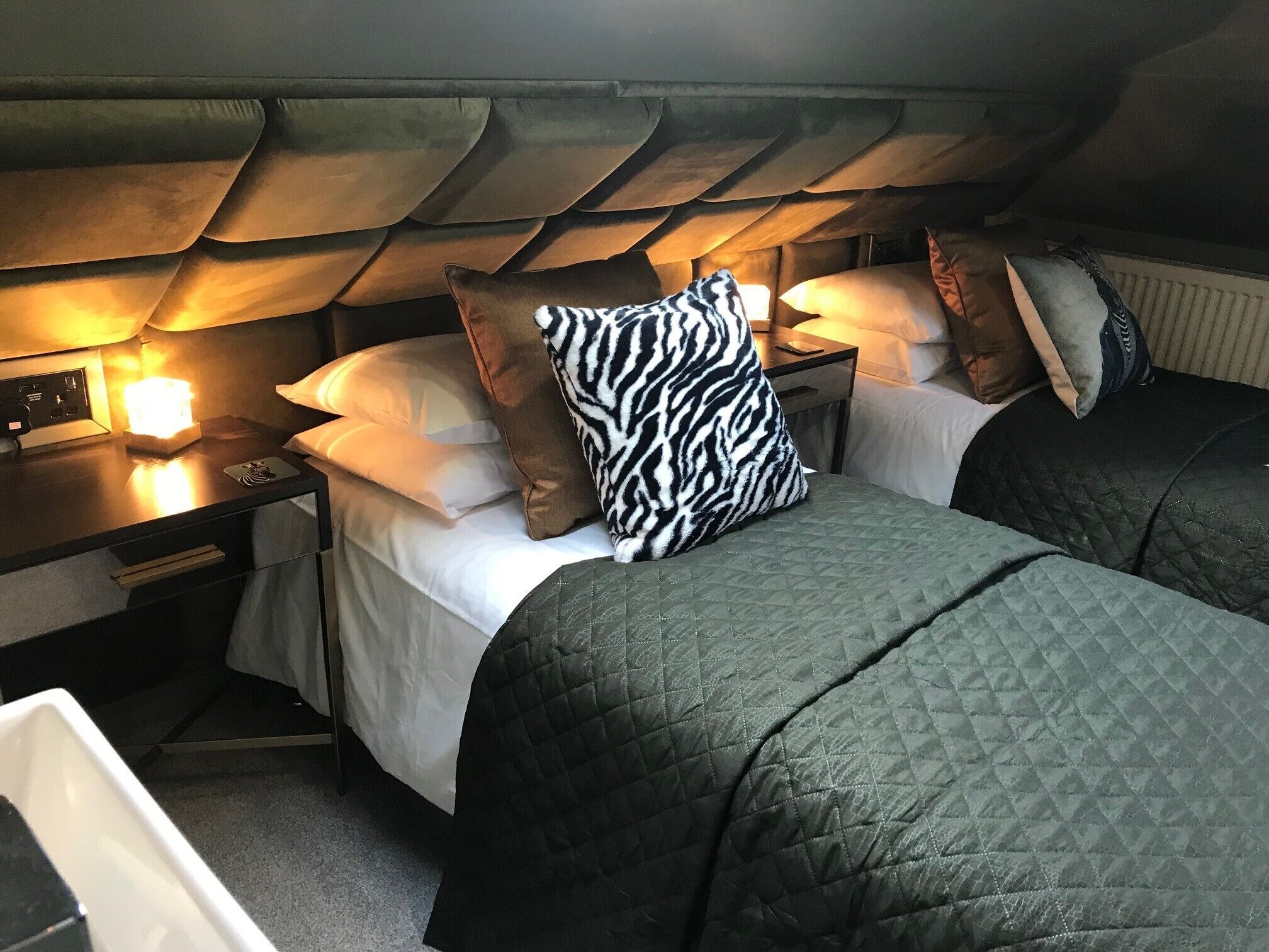 A compact, dimly lit room with two beds, each with a pillow and a bedside table.