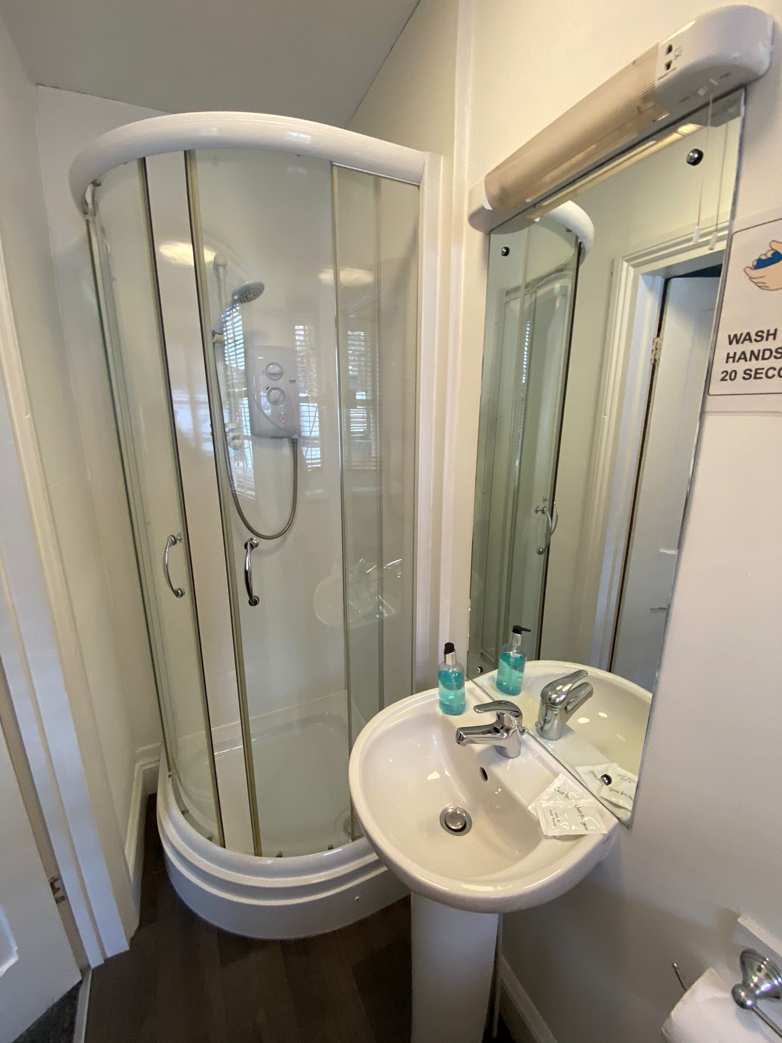 Un baño con ducha de vidrio, lavamanos con espejo y un cartel que indica lavarse las manos durante 20 segundos.