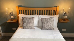 Cama bem arrumada com lençóis brancos e almofadas estampadas, duas mesas de cabeceira com velas e uma obra de arte pendurada na parede.