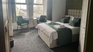 A hotel room with a bed, two chairs, a small table, and a view of the street through large windows.