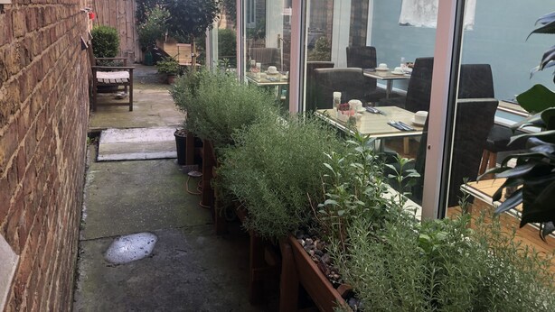 A narrow outdoor patio with a brick wall on one side, a wooden bench, and a table with chairs. There are potted plants along the wall, and a glass door leading to an indoor area with tables and chairs.