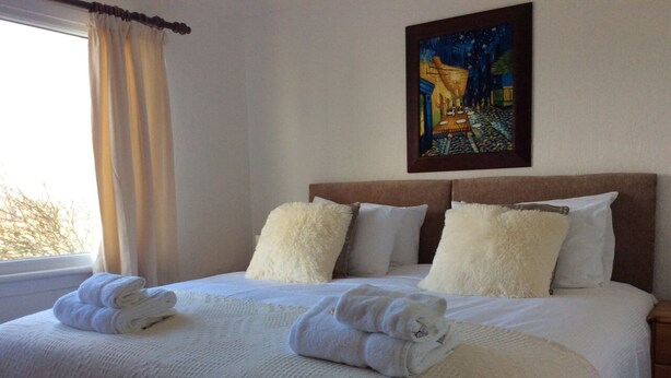 Een netjes opgemaakt bed met witte lakens en kussens, een schilderij aan de muur en een raam met gordijnen.