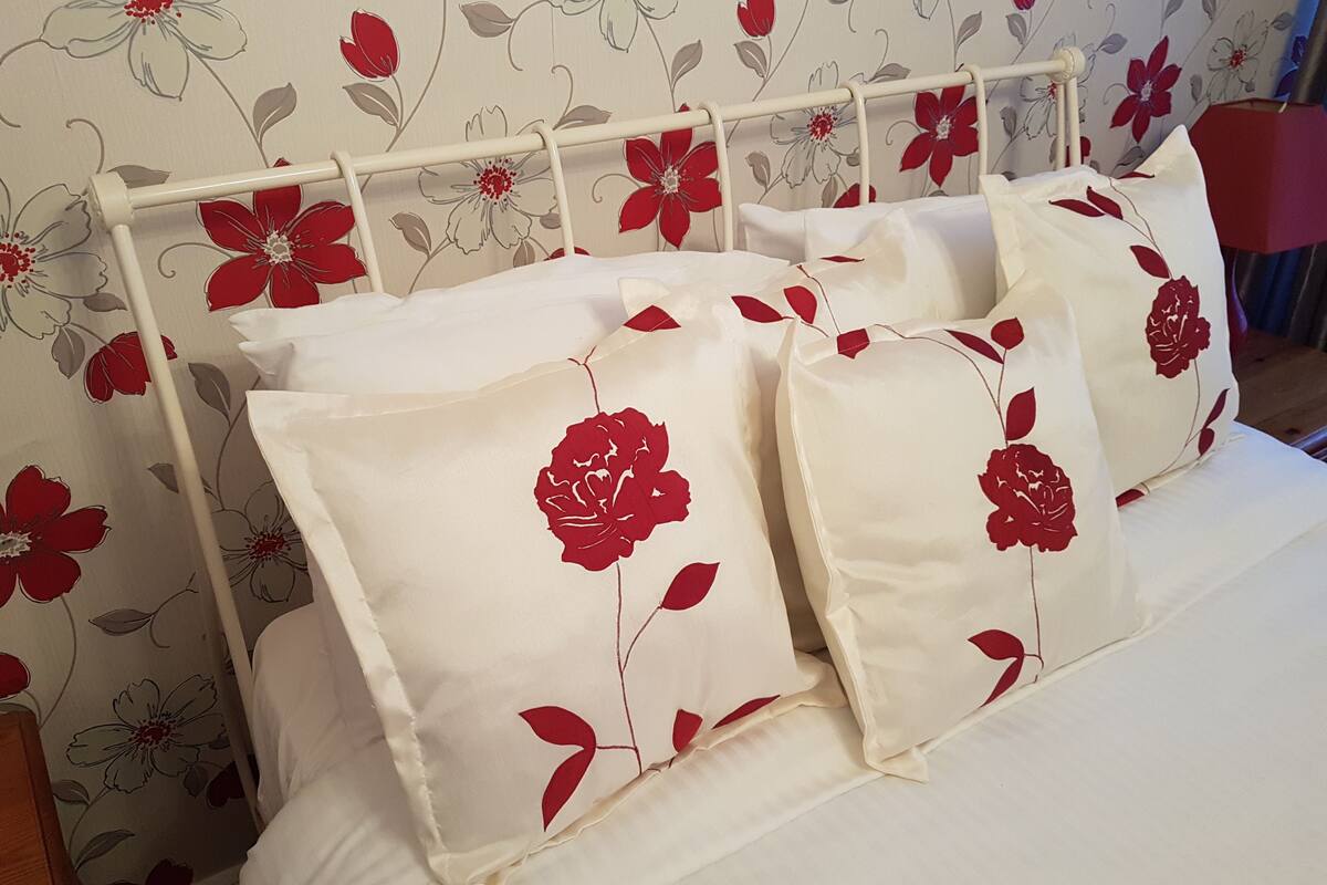 En säng med vita sängkläder och kuddar med röda blommönster, placerad framför en tapet med blommigt motiv.