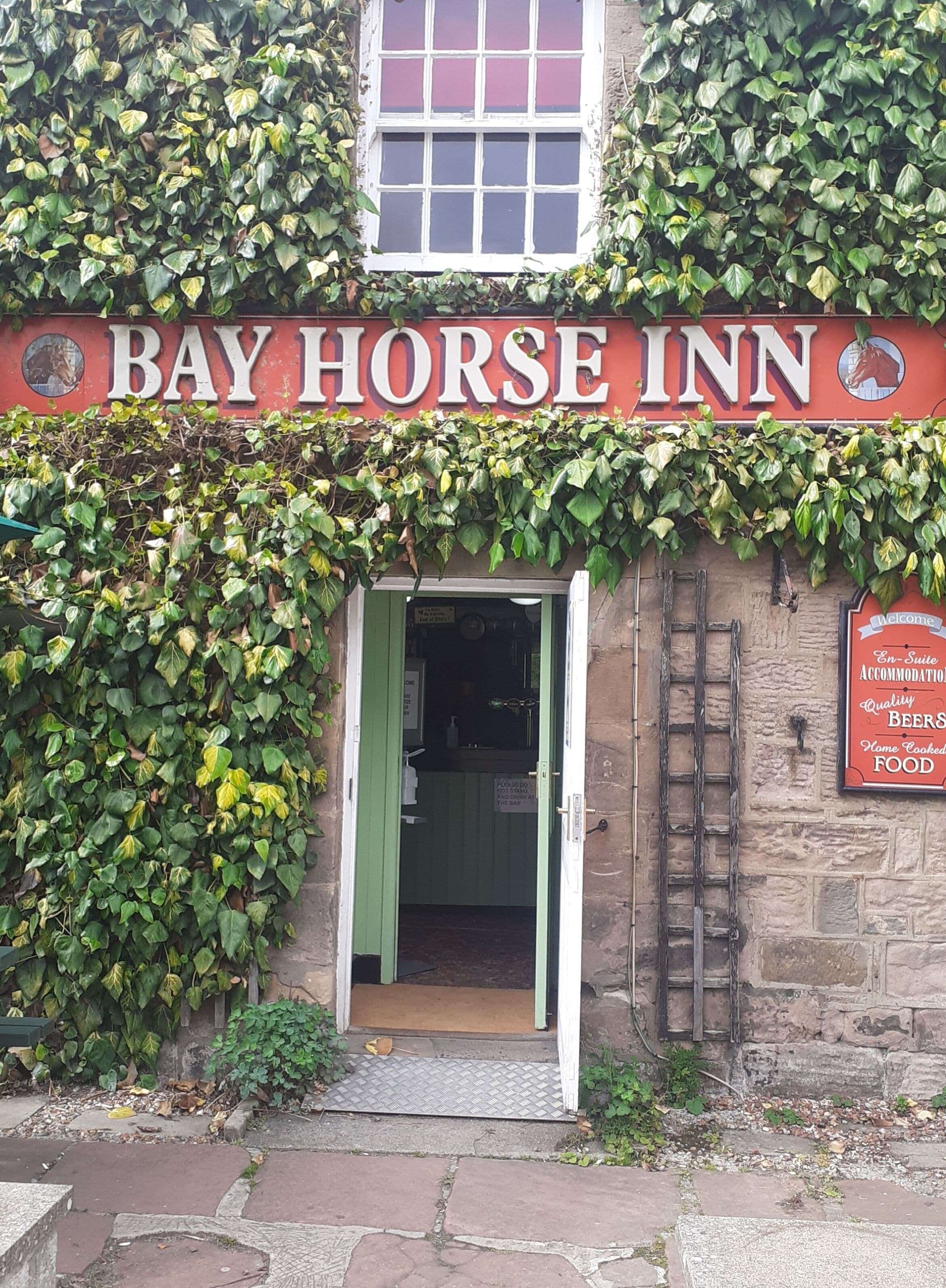 Exterior del Bay Horse Inn, con una puerta verde, muros cubiertos de hiedra y un letrero con el nombre del establecimiento.