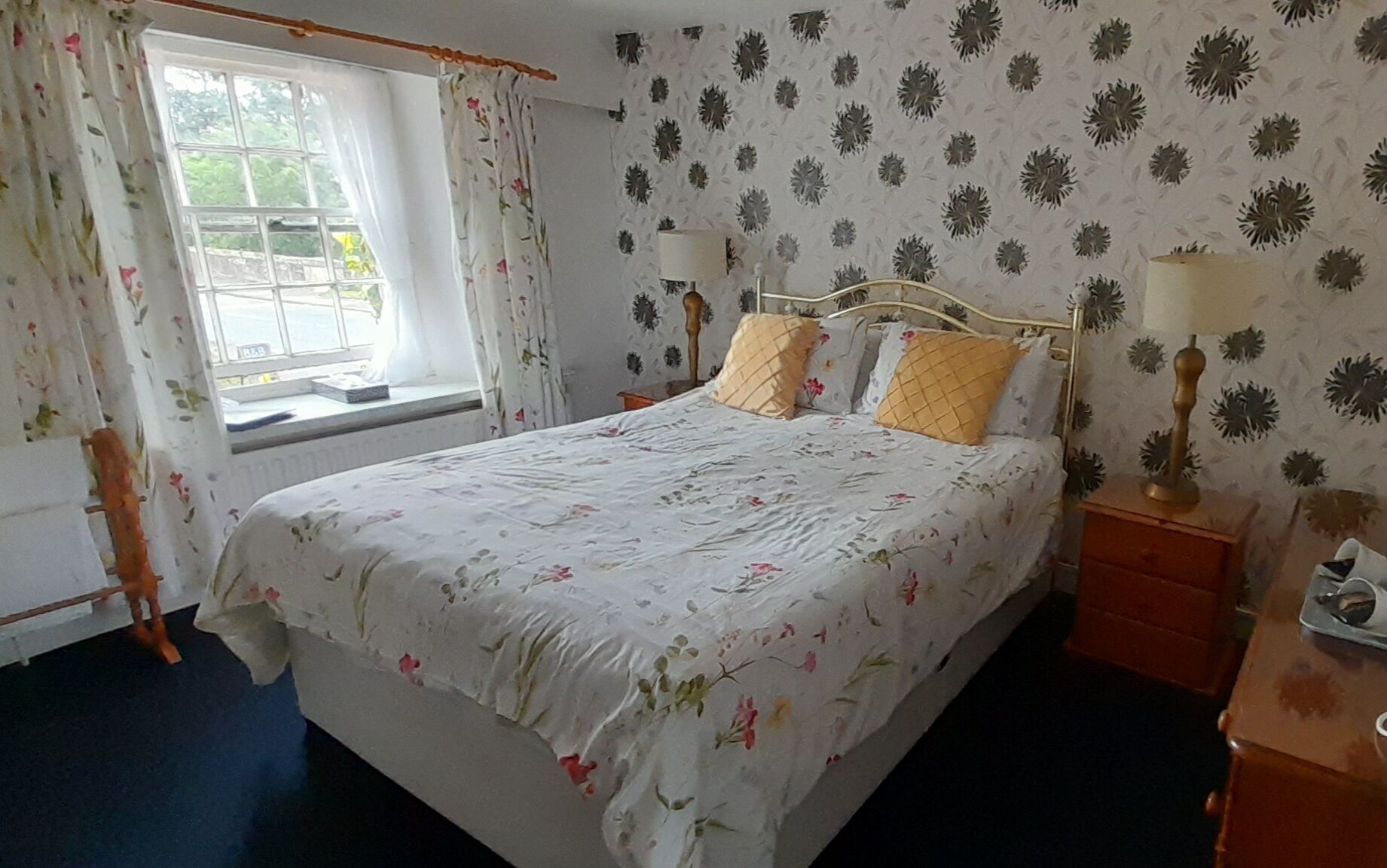 A bedroom with a bed, two bedside tables, a window with curtains, and floral wallpaper.