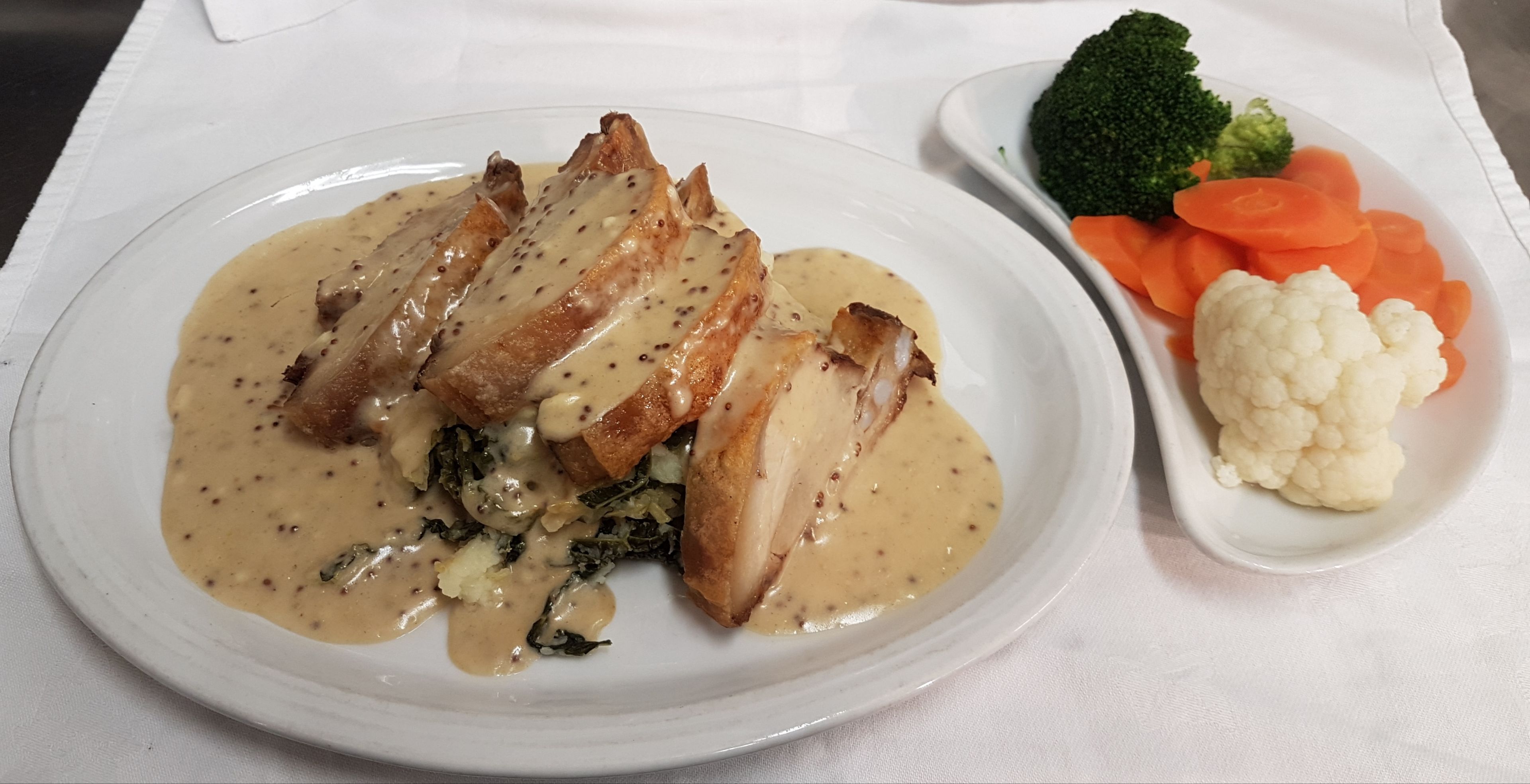Une assiette de nourriture nappée d’une sauce crémeuse, comprenant des tranches de viande et des légumes verts, accompagnée de brocolis, de carottes et de chou-fleur.