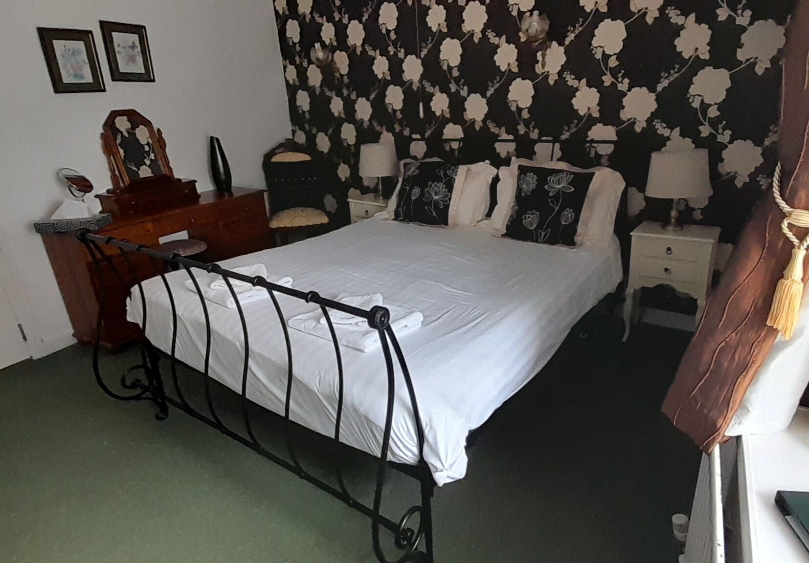 Una camera da letto con una struttura letto in metallo, un comò in legno, un comodino con una lampada e carta da parati a motivi floreali.