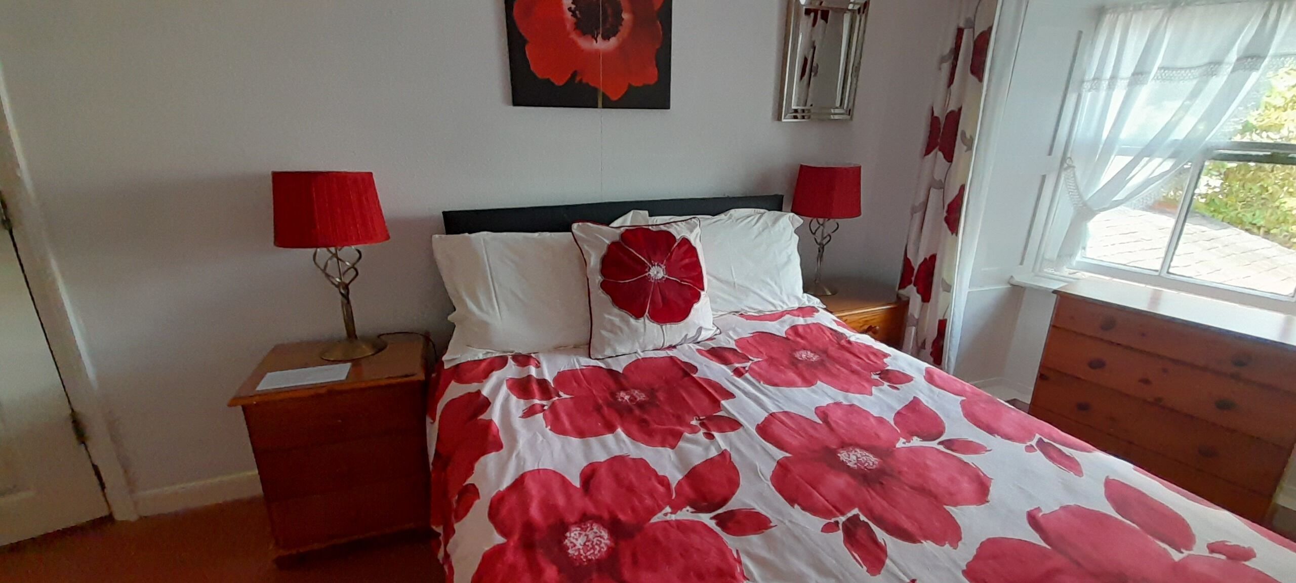A bedroom with a bed, two nightstands, a wooden dresser, a window with curtains, and a wall-mounted lamp.