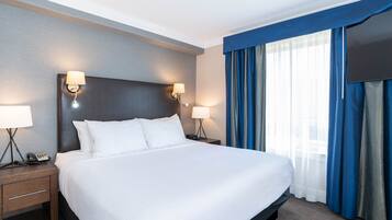 Une chambre d’hôtel avec un grand lit, deux tables de chevet avec des lampes, un téléviseur à écran plat et une fenêtre avec des rideaux bleus.