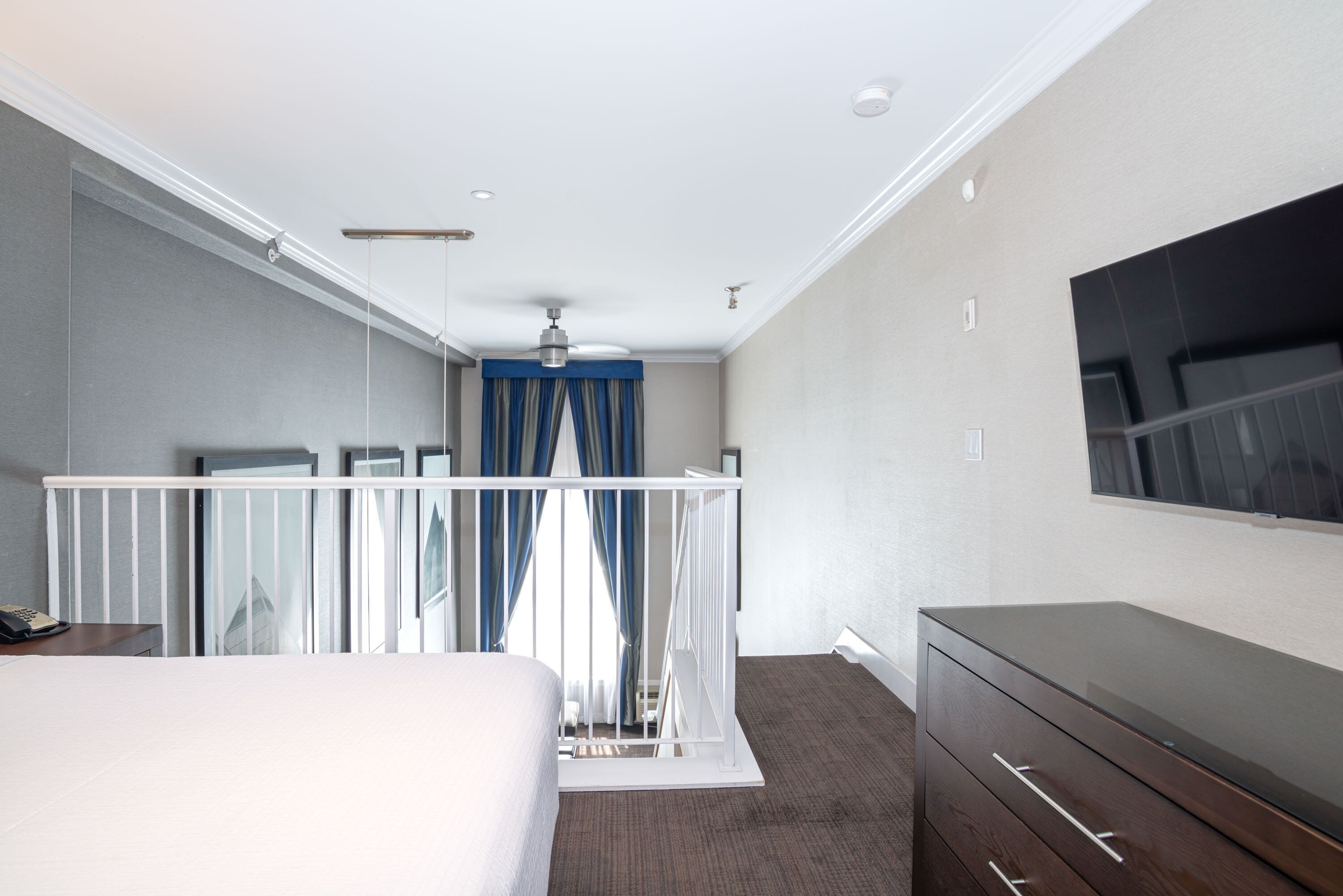 Habitación de hotel con una cama, un aparador, un televisor montado en la pared, un ventilador de techo y una ventana con cortinas.