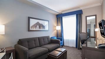 Une chambre d’hôtel équipée d’un canapé, d’un pouf, d’un bureau, d’une chaise, d’une lampe et d’un tableau encadré au mur.