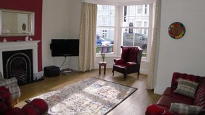 Una sala de estar con chimenea, televisor, un sillón rojo, un sofá rojo y un ventanal con vista a la calle.