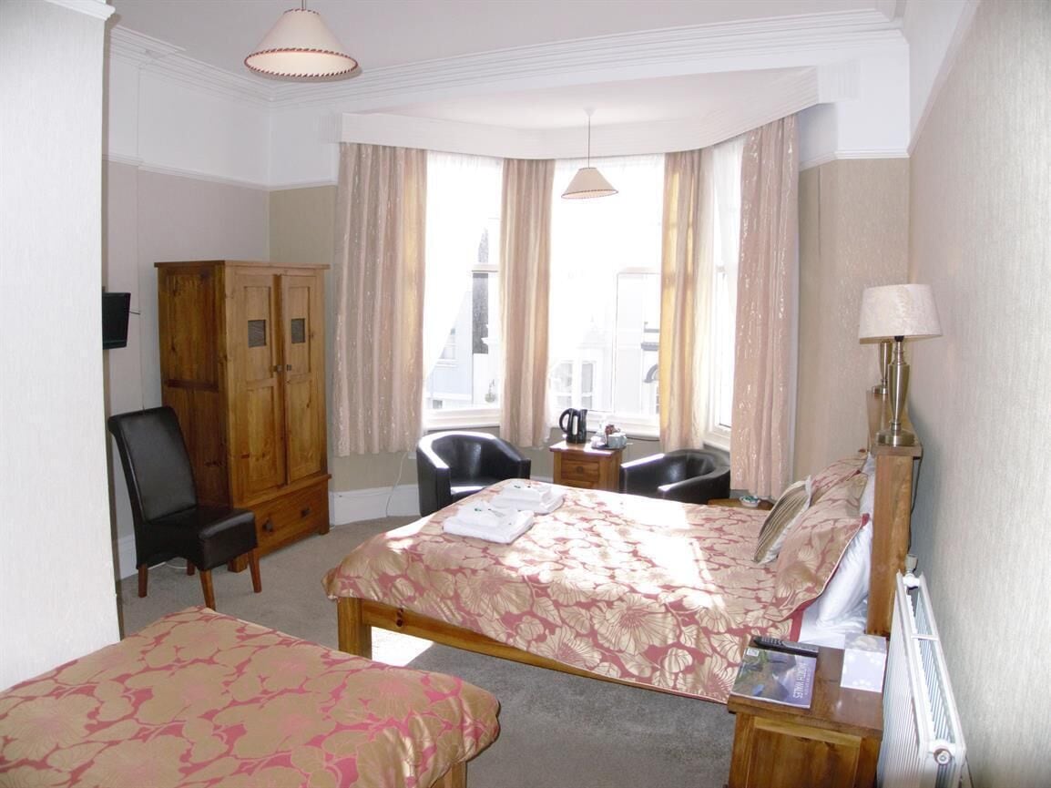 A hotel room with a bed, two chairs, a wooden wardrobe, and a desk with a lamp.