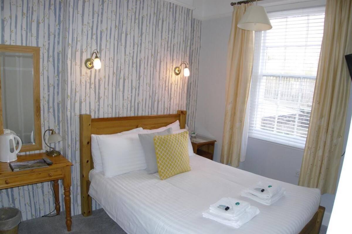 Una cama bien hecha con sábanas blancas, cabecera de madera y almohada amarilla. Hay una mesita de noche con una lámpara y un hervidor. La habitación tiene papel tapiz a rayas y una ventana con cortinas.