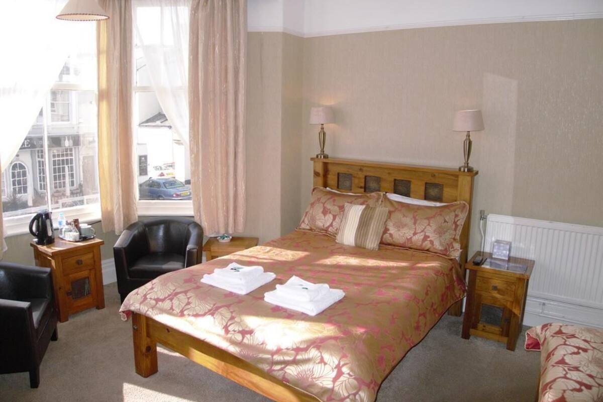 Habitación de hotel con una cama grande, dos mesitas de noche, una silla y vistas a un paisaje urbano a través de amplias ventanas.