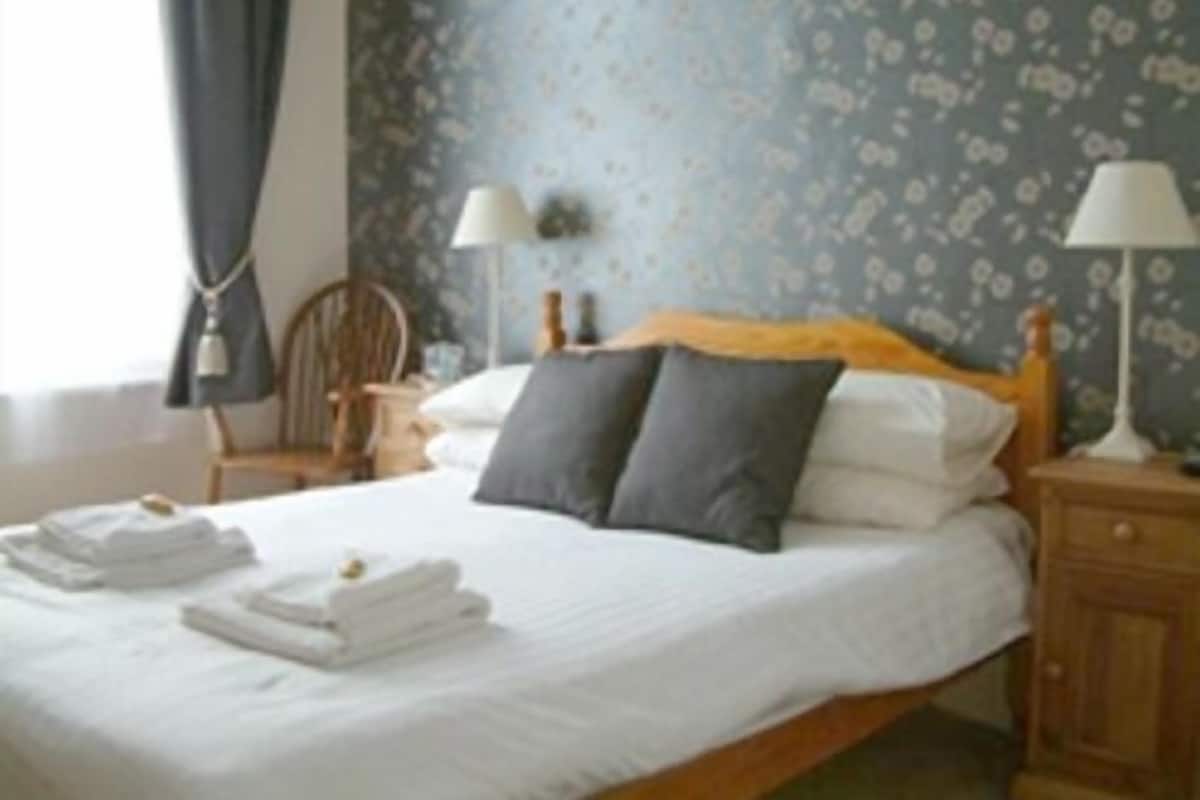 Una cama bien hecha con sábanas blancas y dos almohadas, una mesita de noche de madera con una lámpara, y una silla en una habitación con papel tapiz estampado.