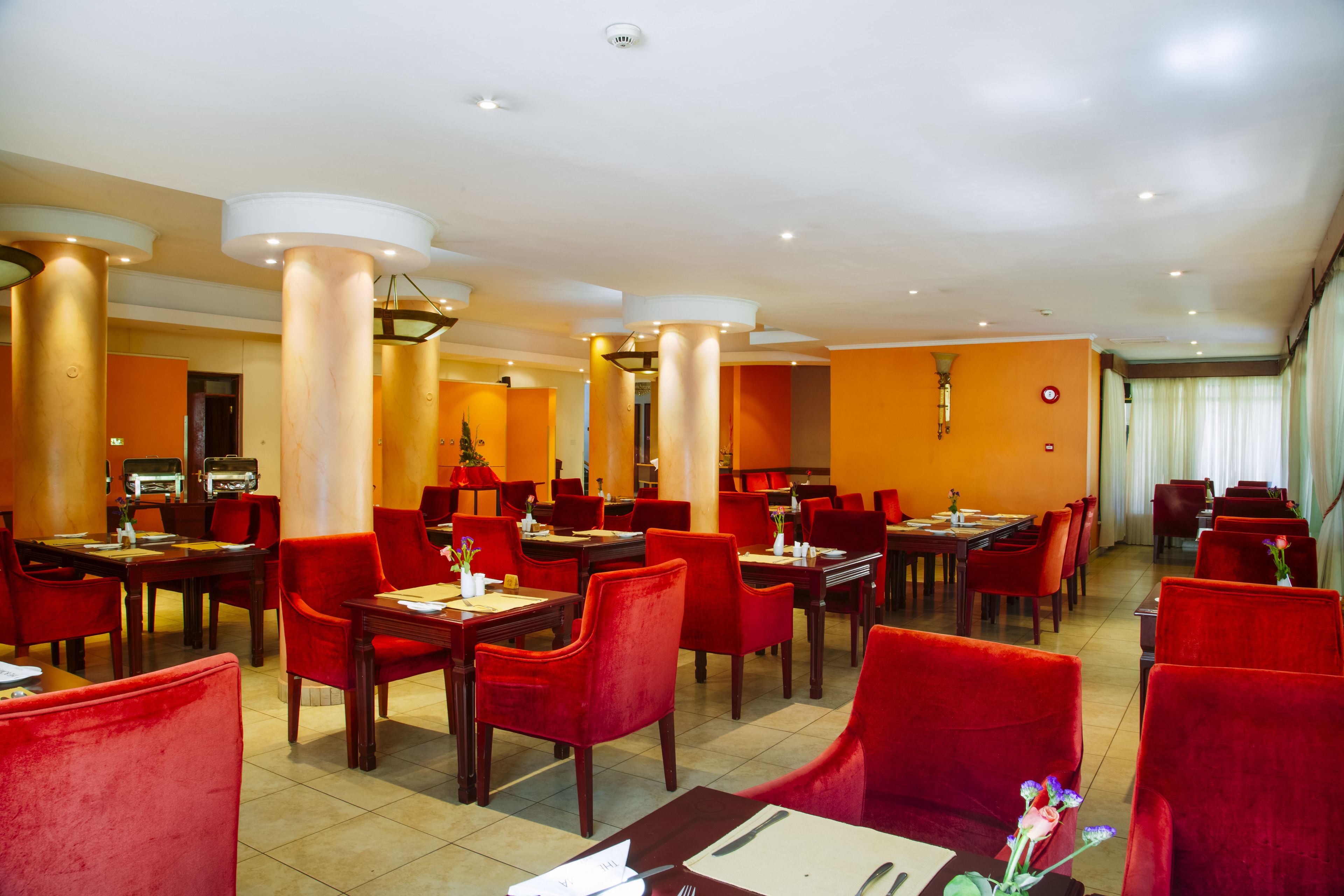 A hotel dining area with red upholstered chairs, wooden tables, and a modern ceiling design.