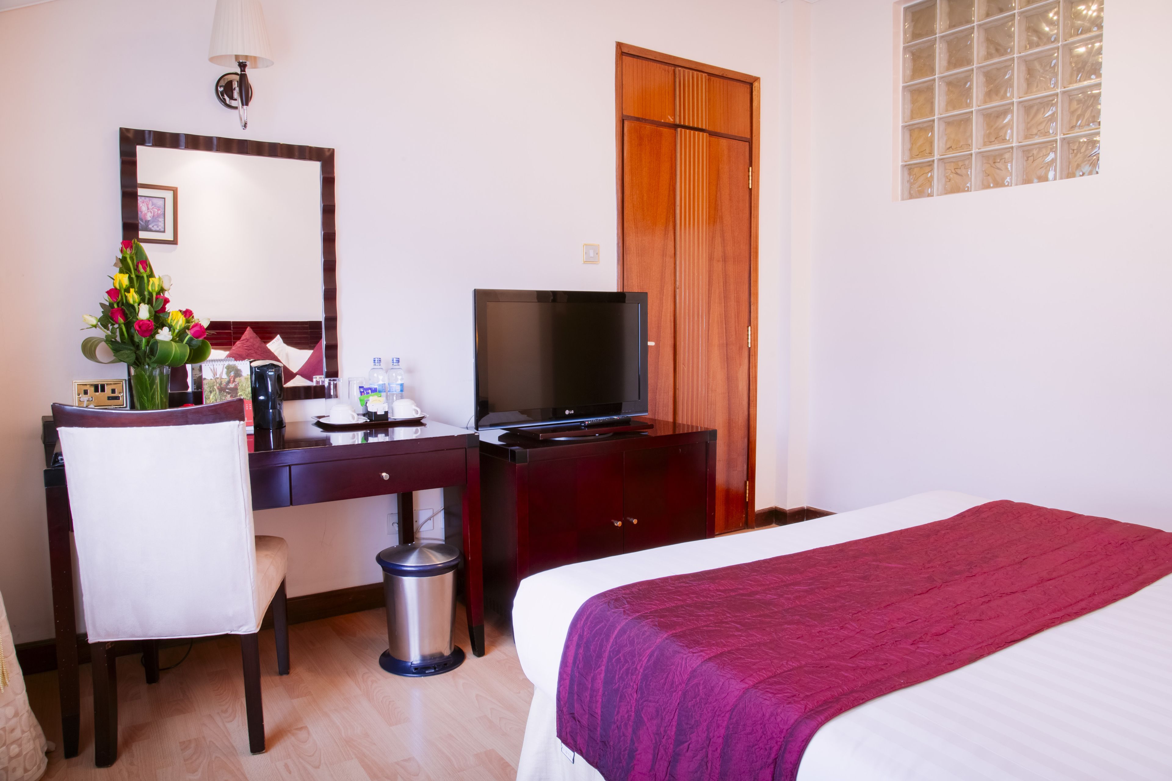 A hotel room with a bed, a desk with a television, a chair, a trash bin, and a wooden wardrobe.