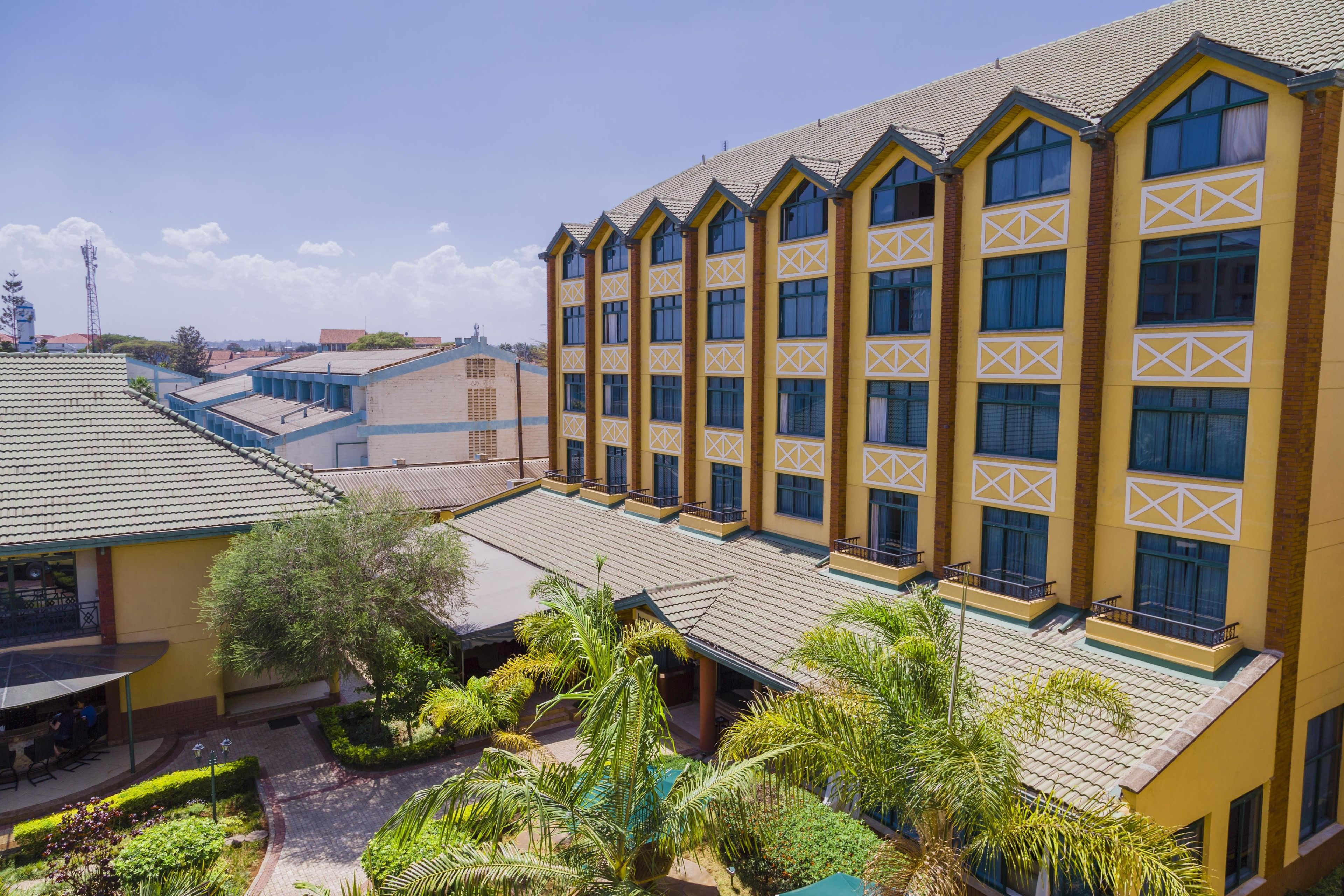 A multi-story hotel with a yellow facade and blue accents, featuring balconies and a landscaped courtyard.