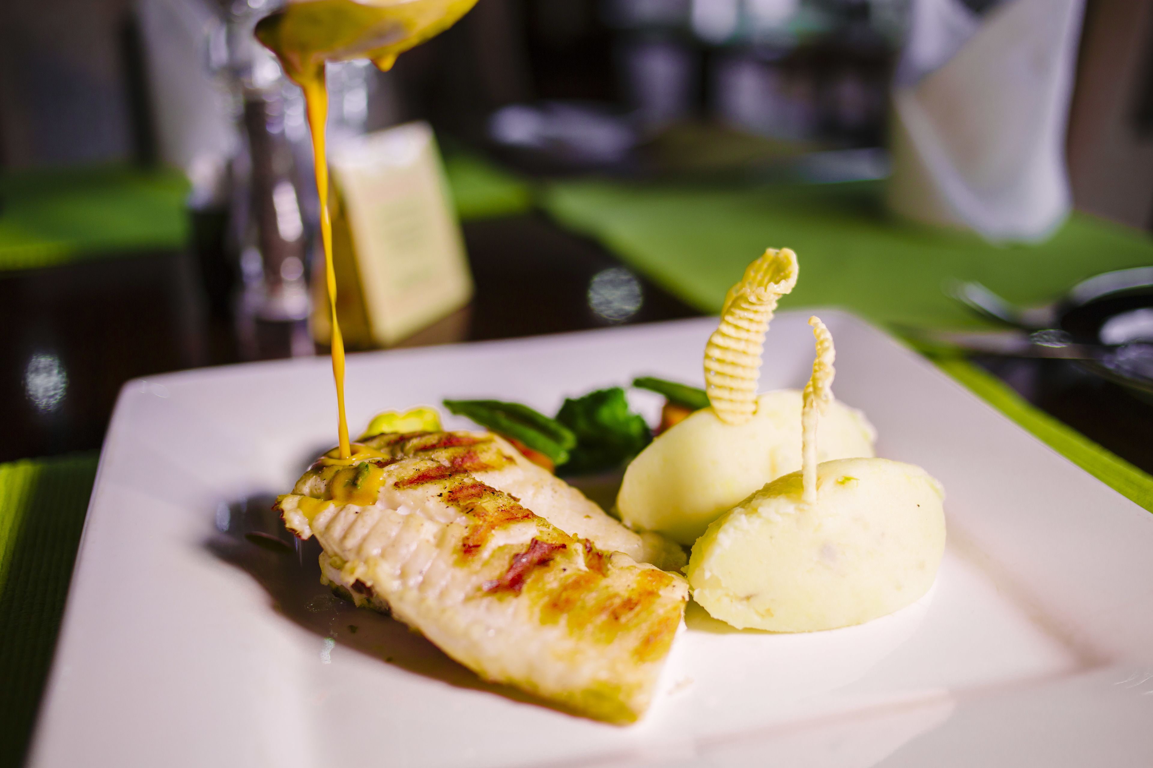 Ein weißer Teller mit gegrilltem Fisch, zwei Knödeln und Soße, die über den Fisch gegossen wird.