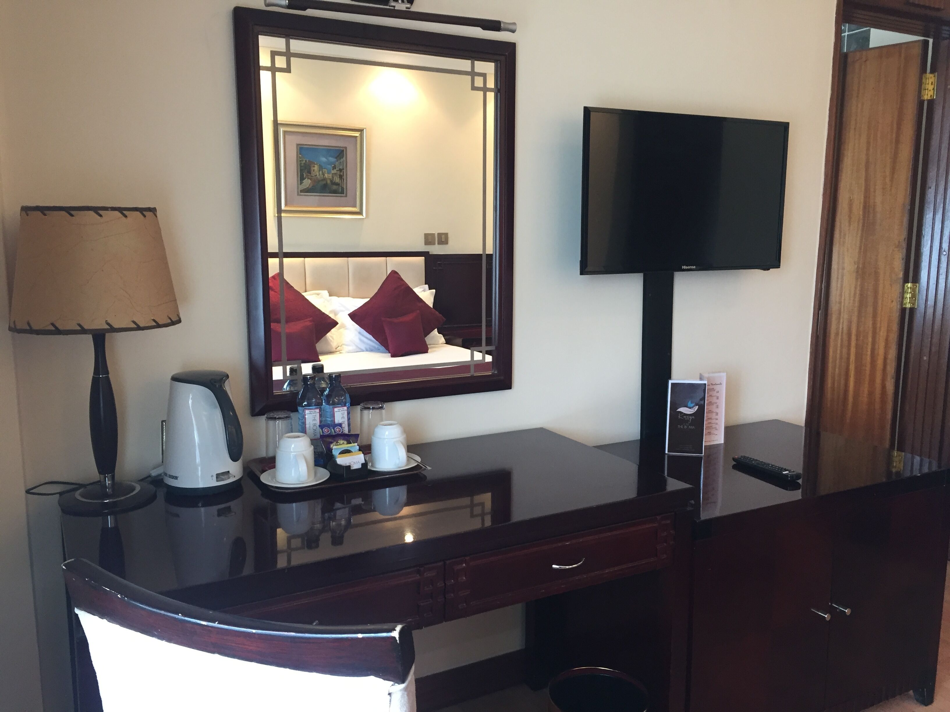 A hotel room with a large mirror, a flat-screen TV, a desk with a coffee maker, and a bed with red pillows.