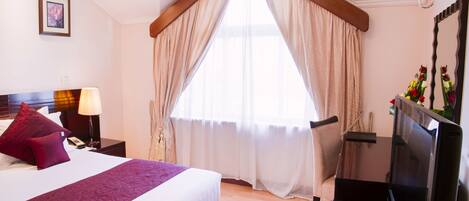 Una habitación de hotel con una cama grande, un escritorio con televisor y un ventanal con cortinas.