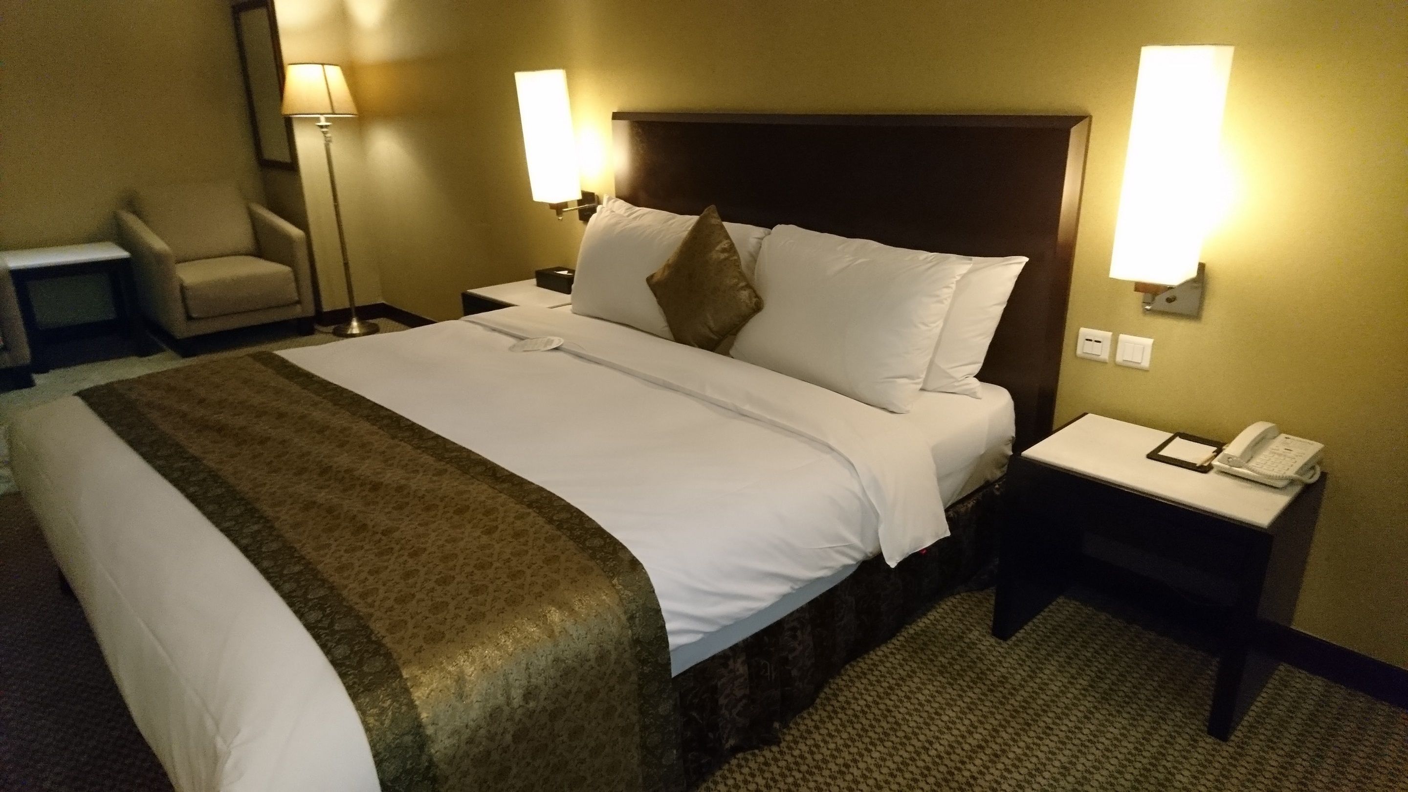 A hotel room with a large bed, two bedside lamps, a chair, a bedside table with a phone, and a carpeted floor.