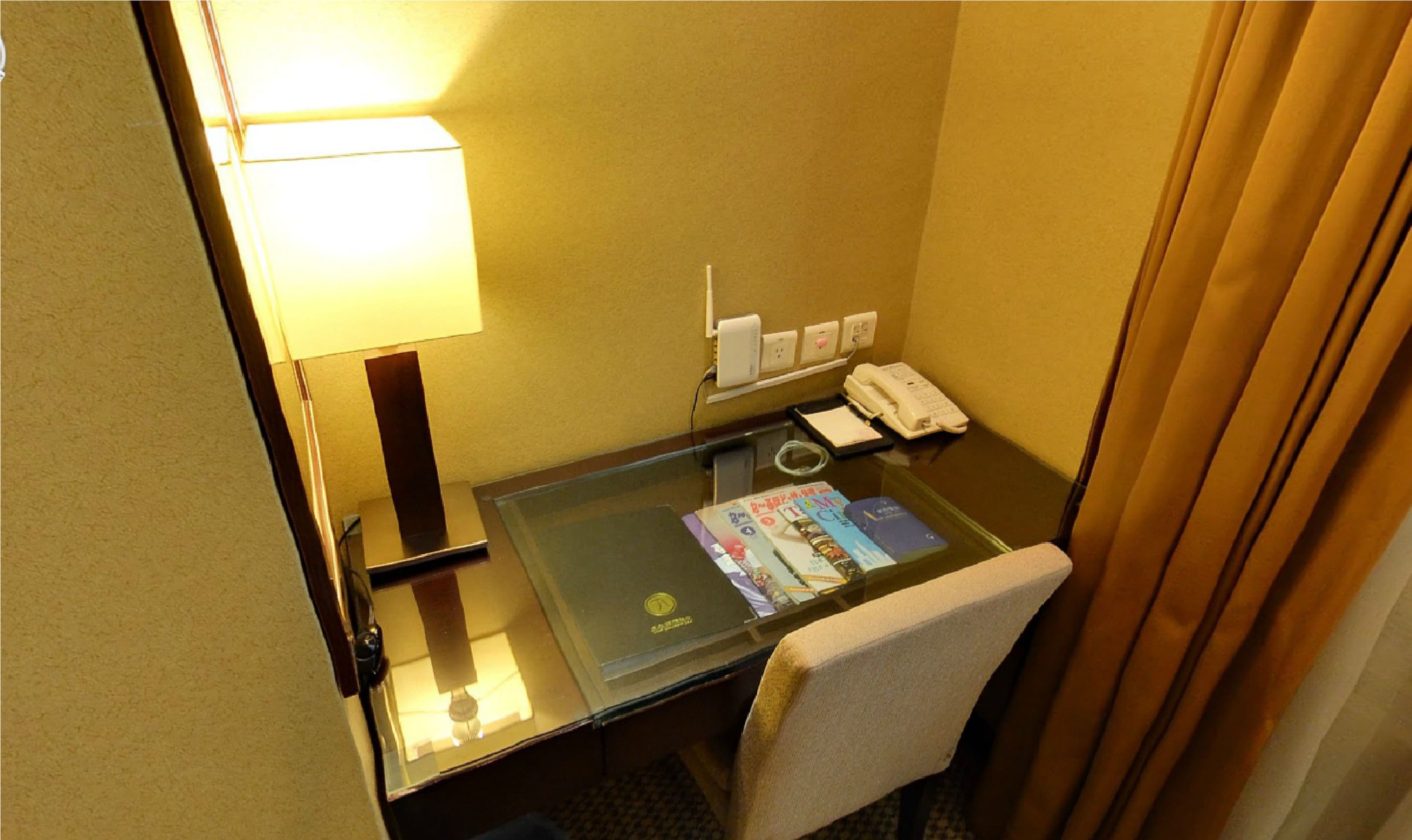 A hotel room with a glass desk, a lamp, a telephone, and a chair.
