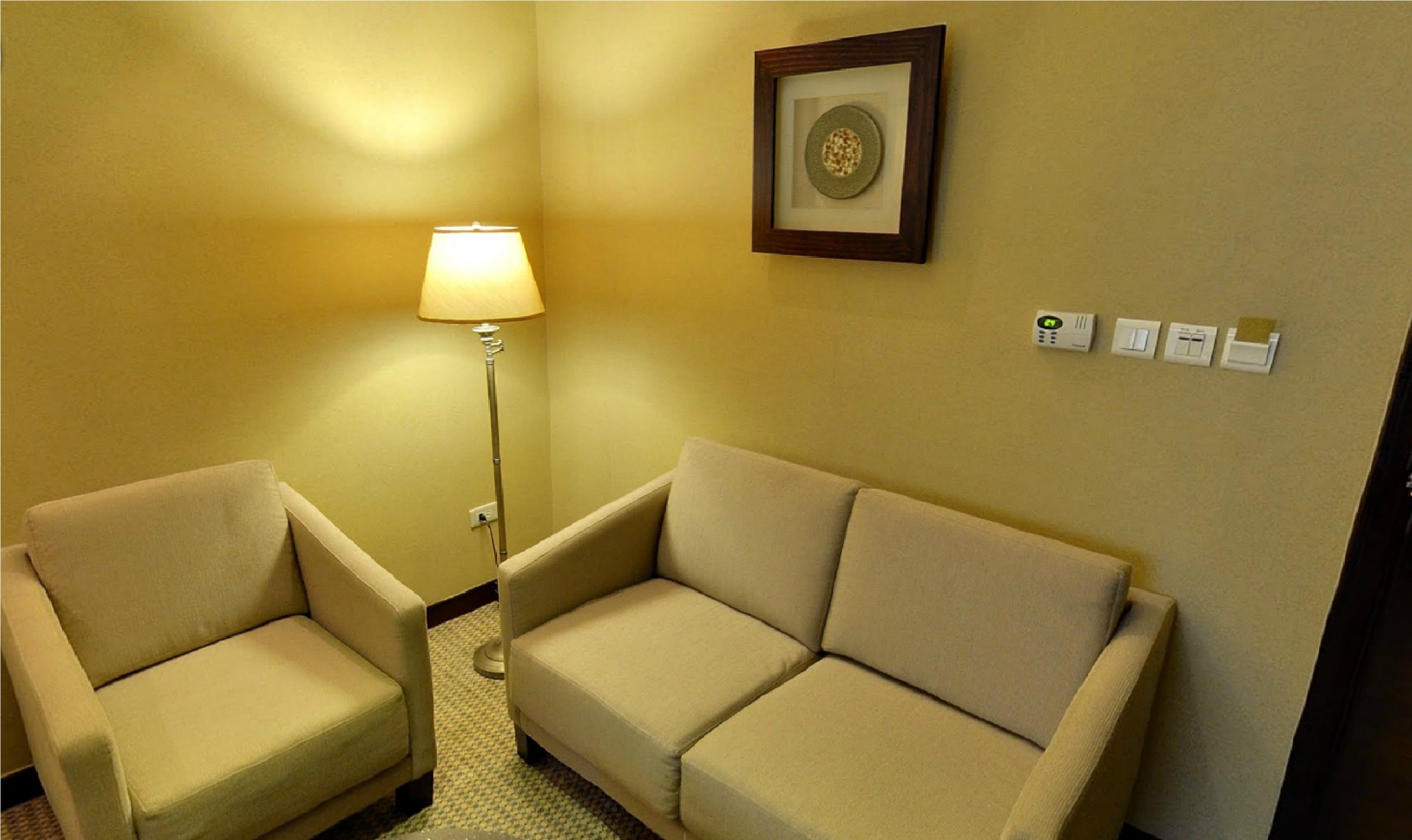 A hotel room with two beige armchairs, a floor lamp, a framed artwork on the wall, and a thermostat.