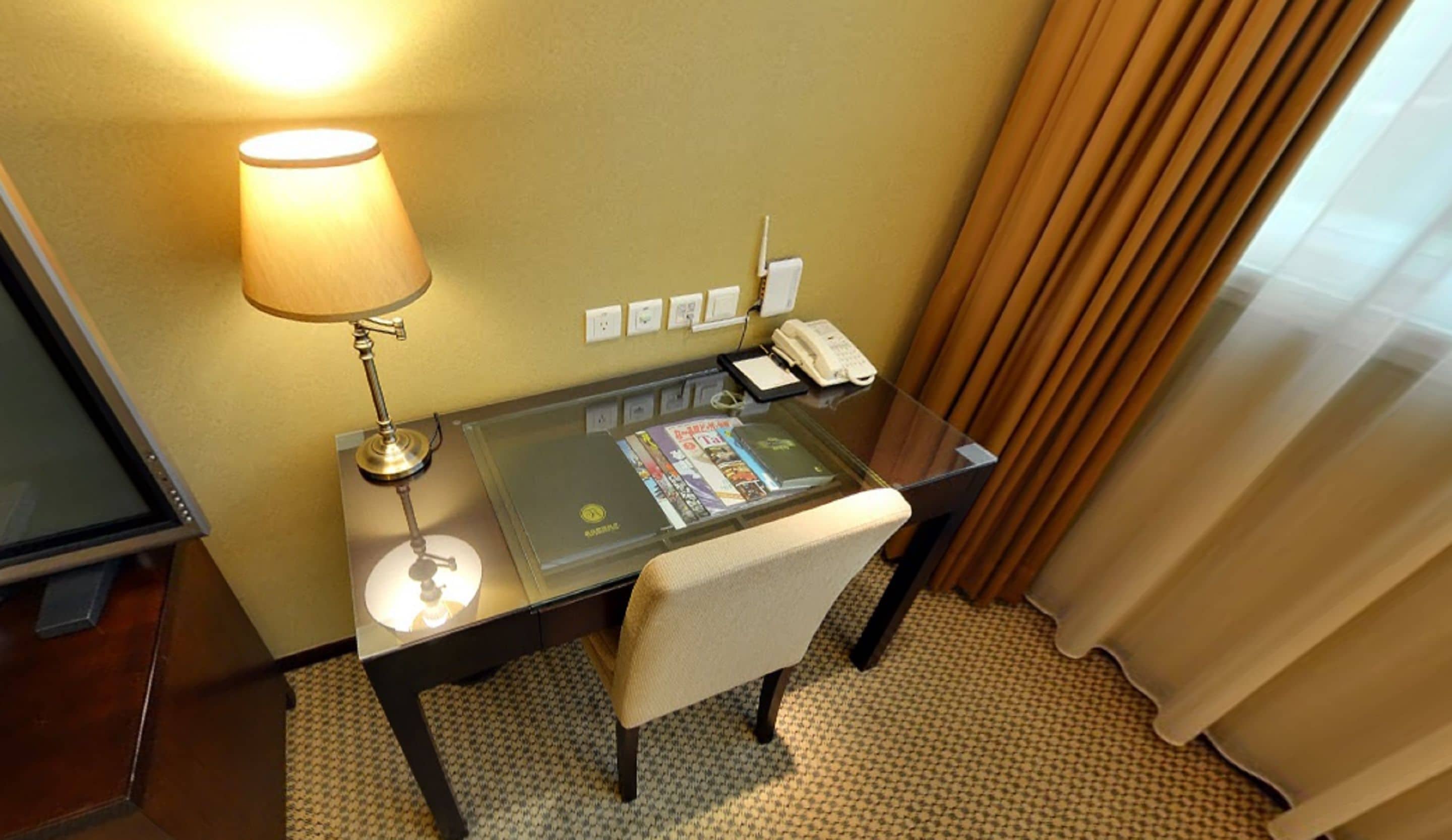A hotel room with a glass desk, a lamp, a telephone, and a chair.