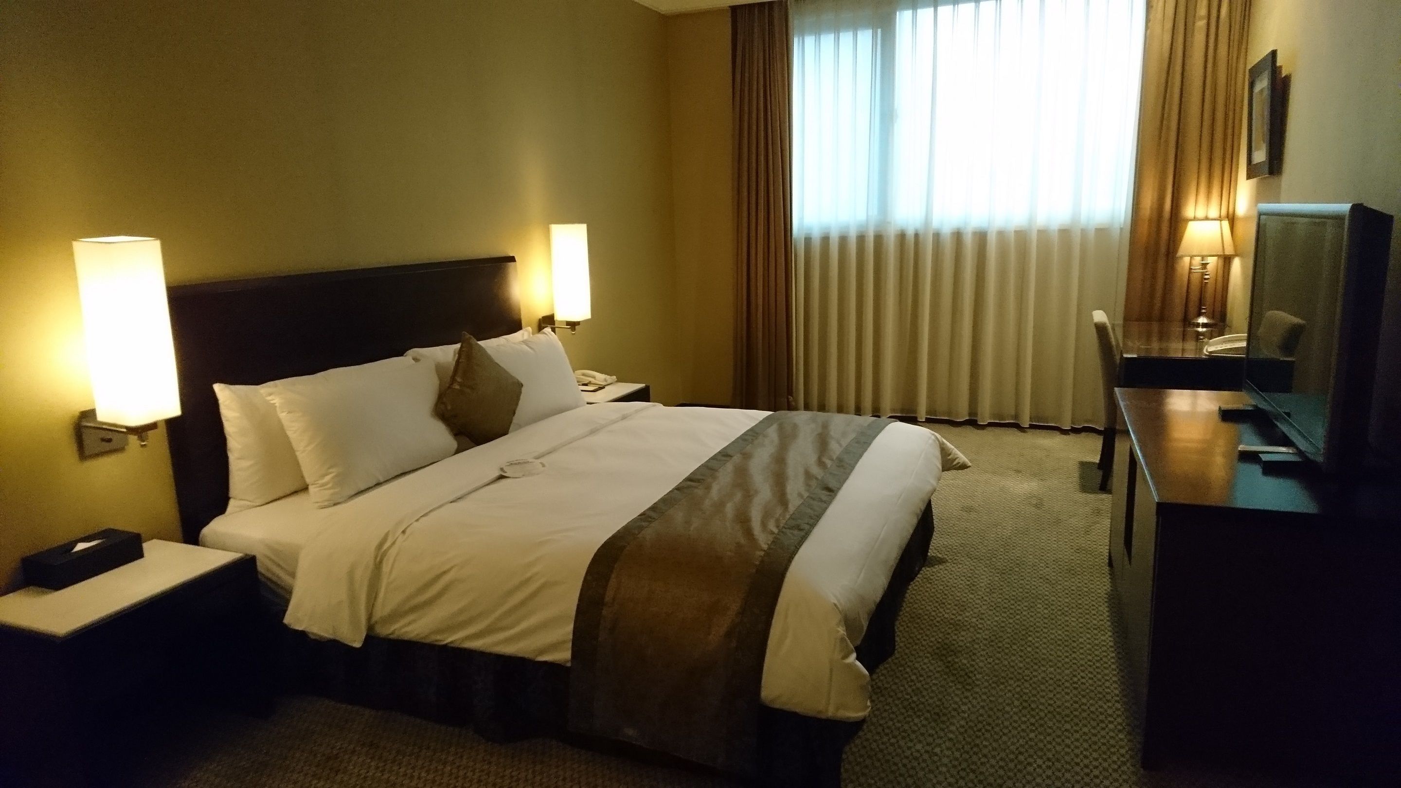 A hotel room with a large bed, two bedside lamps, a desk with a chair, and a television.