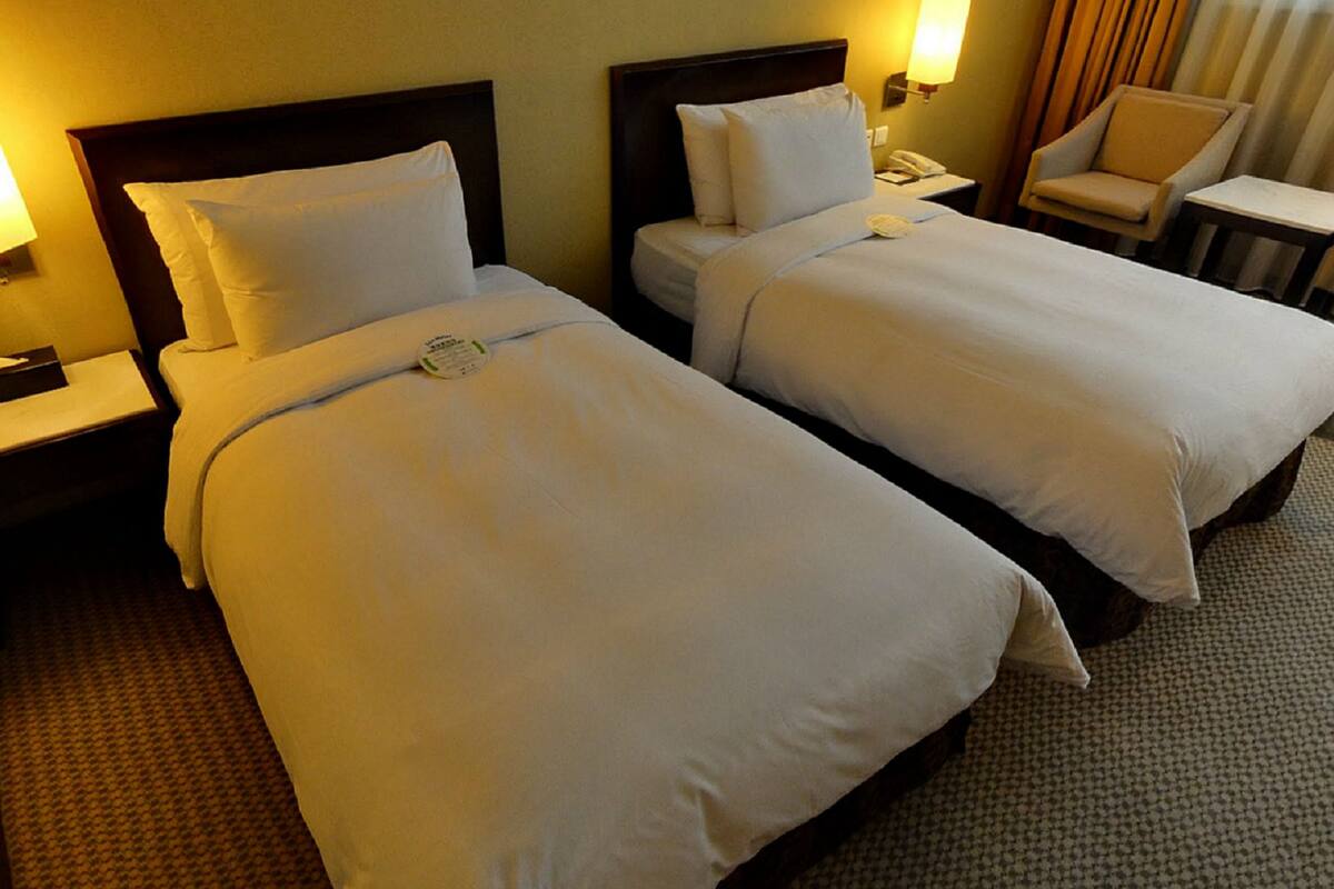 Une chambre d’hôtel avec deux lits, chacun doté d’une tête de lit en bois, de draps blancs et d’une table de chevet avec une lampe. On y trouve également une chaise et une petite table dans un coin.