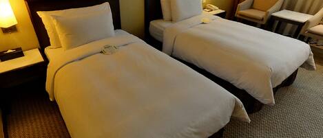 A hotel room with two beds, each with a wooden headboard, white bedding, and a bedside table with a lamp. There is a chair and a small table in the corner.