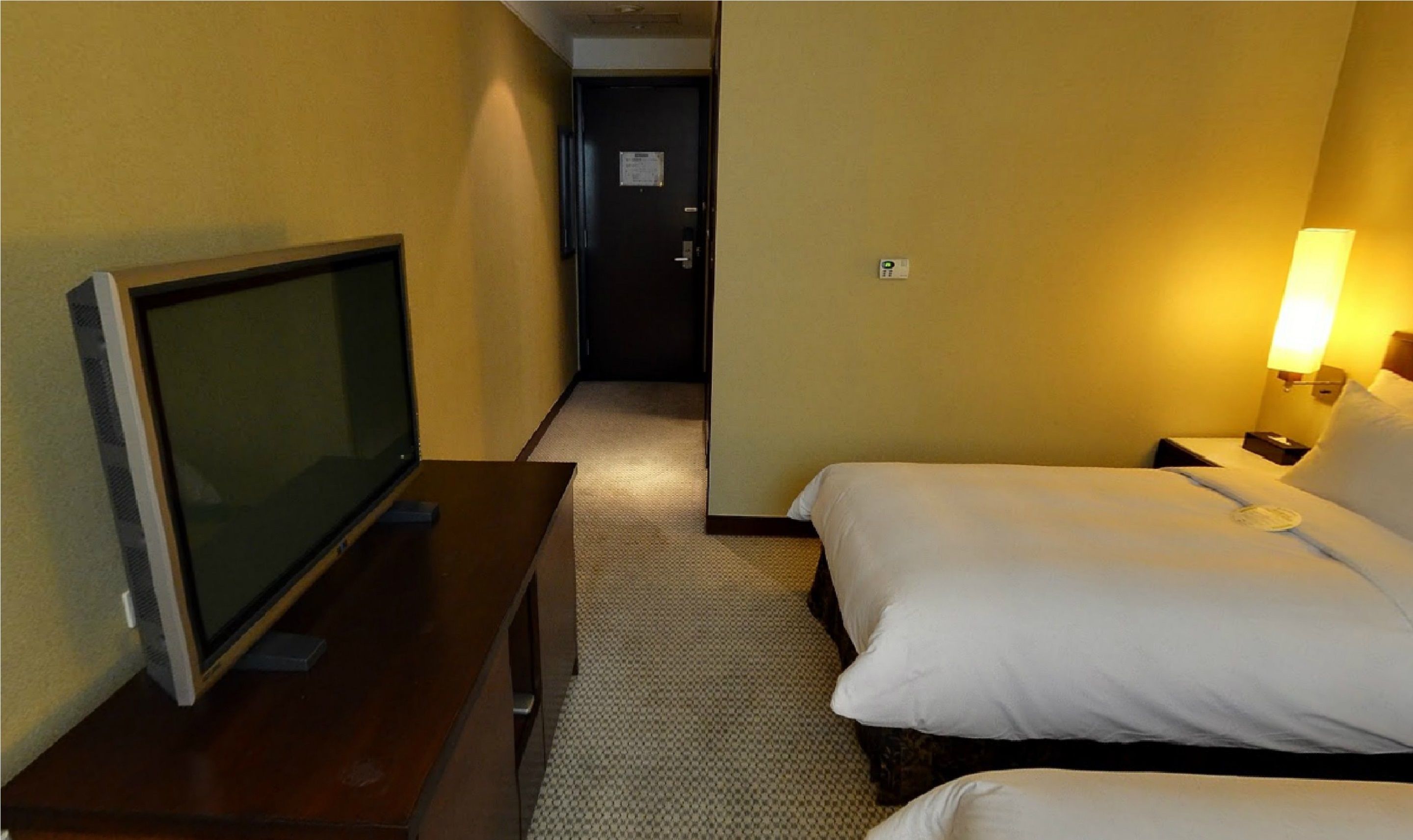A hotel room with a television on a desk, a bed with white bedding, and a nightstand with a lamp.