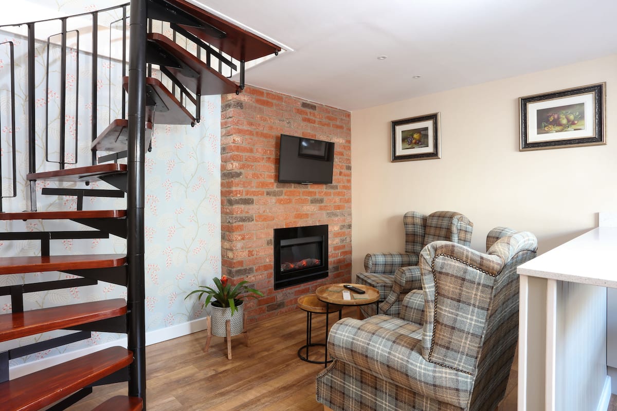 A living room with a brick fireplace, a wooden staircase, and plaid furniture.