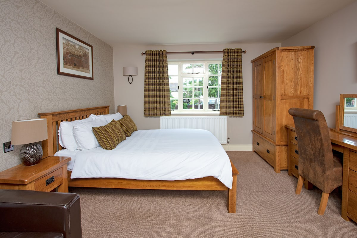 A bedroom with a wooden bed, a wooden desk, a chair, a lamp, and a window with curtains.